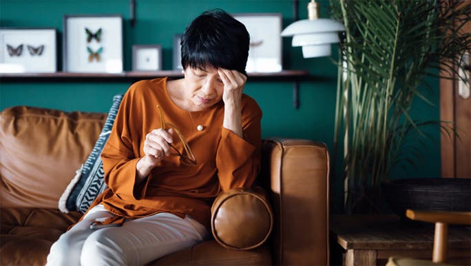 An Asian woman is sitting on a brown leather sofa with her head in her hand